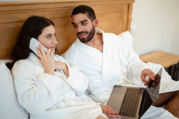 Σύζυγος που δείχνει φορητό υπολογιστή προς σύζυγο ενώ μιλάει στο τηλέφωνο κάθεται στο κρεβάτι εσωτερικά, φορώντας λευκά μπουρνούζια. Ανεξάρτητη και κινητή αντίληψη επικοινωνίας. Επιλογή εστίασης - Φωτογραφία, εικόνα