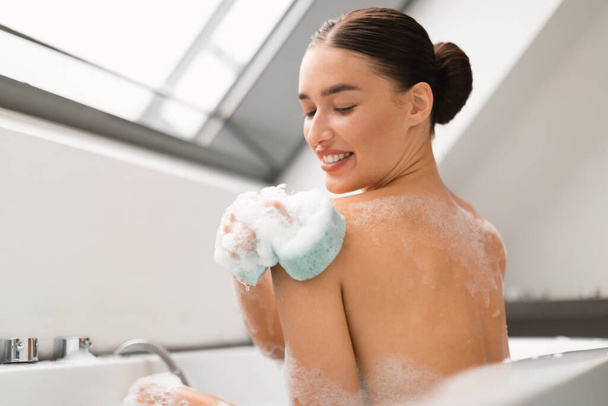 Женщина принимает ванну с пеной и мытье тела с помощью губки трения плеча сидя в ванной в современной ванной комнате в помещении. Красота и косметика. Селективный фокус - Фото, изображение