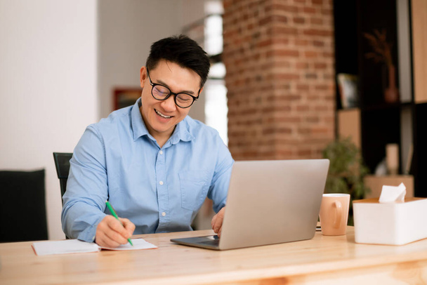 Boldog ázsiai férfi jegyzetel, elemzi a munkahelyi befektetésekre vonatkozó adatokat laptoppal az otthoni irodai belső térben. Munka, üzleti és startup tervezés, ötletbörze távirányítással - Fotó, kép