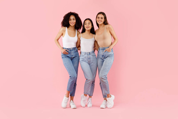 Felice tre ragazze multirazziali che abbracciano e sorridono alla macchina fotografica, in posa in studio su sfondo rosa, full body lenght shot. Concetto di amicizia femminile - Foto, immagini