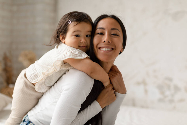Веселая азиатская мама обнимается и обнимается с маленькой дочкой, улыбаясь в камеру, сидя дома на кровати. Мама развлекается, играя с маленькой девочкой в спальне. Моменты материнства - Фото, изображение