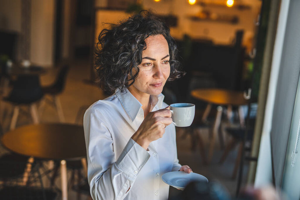 One woman stand at the window at home or office businesswoman entrepreneur wear white shirt having cup of coffee happy smile real people copy space morning routine mature caucasian female - Foto, afbeelding
