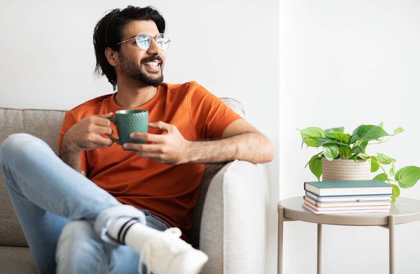 Glad millennial arab guy in casual, glasses with beard sits on sofa with cup of hot drink in living room interior, look at free space. Lifestyle, tea and relax from work, coffee break, ad and offer - Фото, зображення