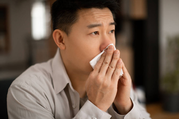 Griep. Zieke Aziatische man van middelbare leeftijd die niest met een loopneus, thuis zit en ruimte kopieert. Man die last heeft van koude en hoge koorts, zich niet lekker voelt. Begrip virusziekte - Foto, afbeelding