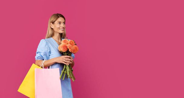 Smiling middle aged european blonde female with packages with purchases and bouquet of flowers enjoys shopping isolated on pink studio background, panorama. Sale and holiday celebration - Foto, immagini