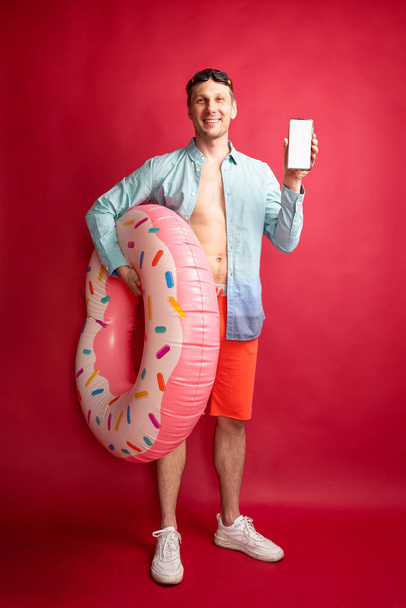 Young smiling happy tourist man in beach shirt hold inflatable ring hold in hand use mobile cell phone isolated on red background studio portrait. Summer vacation sea rest sun tan concept. - Foto, Bild