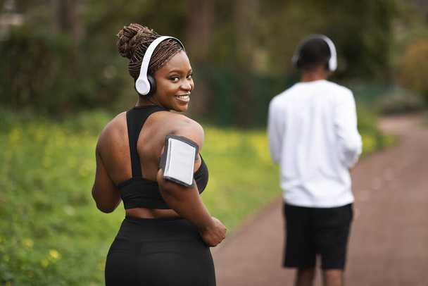 Smiling millennial african american couple in sportswear, wireless headphones running with smartphone outdoor, back. Weight loss with music, jogging together, sport training, body care - Fotoğraf, Görsel