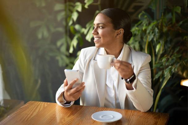 Sorridente donna d'affari millenaria afroamericana in abito bianco guardare fuori finestra con tazza di bere chiacchierando allo smartphone in caffè con piante verdi interni. App per aziende moderne, lavoro, stile di vita - Foto, immagini