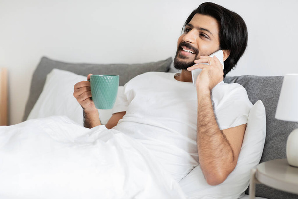 Lächelnder Millennial-Araber mit Bart liegt mit einer Tasse Kaffeeanruf per Smartphone im Schlafzimmer. Klatsch und Tratsch, gute Nachrichten, guten Morgen und Tee, Entspannung, Kommunikation zu Hause - Foto, Bild