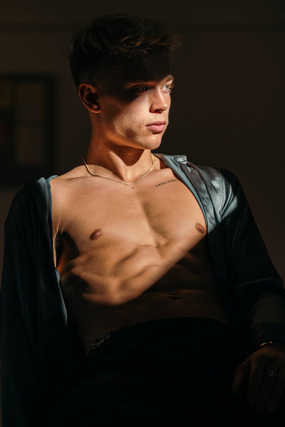 A muscular man in a satin shirt looking to the side on a dark background with falling sunlight - Photo, Image