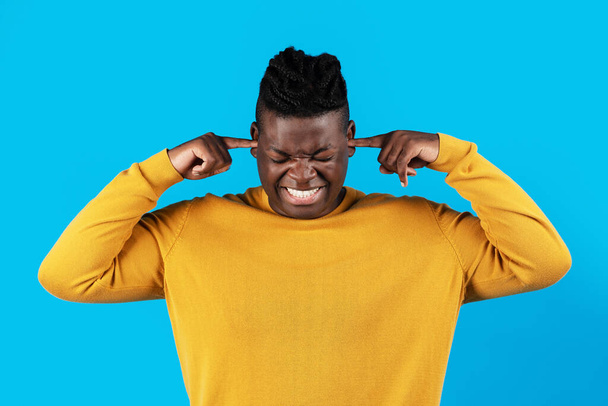 Annoyed young african american man plugging ears with fingers and frowning, angry millennial black male doesnt want to hear something, standing isolated over blue studio background, copy space - Foto, afbeelding