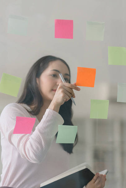 A professional millennial Asian businesswoman putting colorful sticky notes on a glass window in a creative office. Business planning and brainstorming strategy concept - Foto, imagen