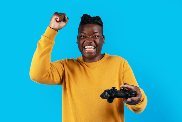 Videojáték-koncepció. Eufórikus afro-amerikai férfi Joystick ünneplő játék győzelem, boldog fekete srác felemeli öklét, és felkiáltva izgalommal, álló elszigetelt kék háttér - Fotó, kép