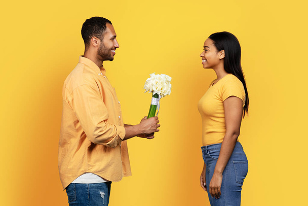 Cheerful millennial african american guy gives bouquet to woman, congratulation happy birthday, anniversary isolated on yellow studio background. Relationship, romance love and date - Foto, Imagem