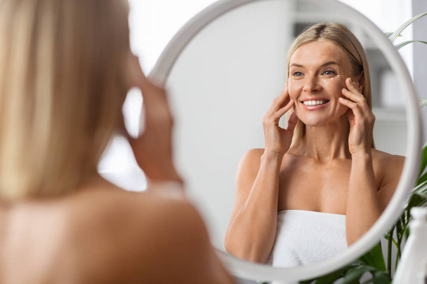 Anti-Aging Cosmetics. Beautiful Middle Aged Woman Applying Eye Cream While Standing Near Mirror At Home, Attractive Mature Lady Touching Her Face, Enjoying Daily Beauty Routine, Selective Focus - Foto, imagen