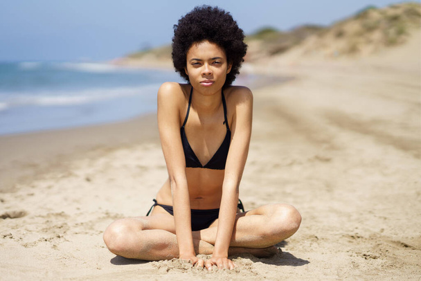 Full body of slim African American female traveler, in swimwear looking at camera with serious face, while sitting on sandy coast near sea during summer vacation - Photo, Image