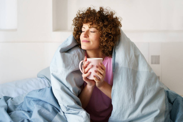 Relaxed curly woman with closed eyes wrapped in blanket while holding mug of hot beverage on bed in weekend - Photo, Image
