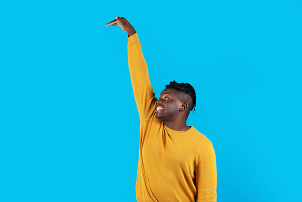 Cheerful African American Guy Measuring High Invisible Object With Hand While Standing Isolated Over Blue Studio Background, Young Black Man Looking Up And Checking Hight, Copy Space - Φωτογραφία, εικόνα