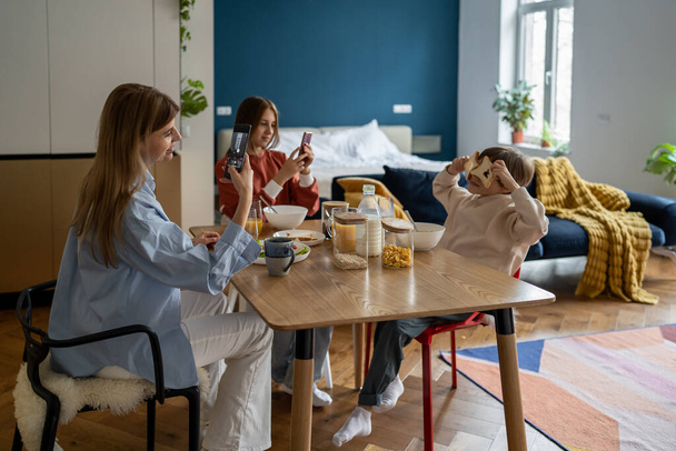 Family breakfast with addiction from smartphone. Pleased mom takes pictures as kid son fooling around with bread toast while daughter look at screen cellphone at kitchen table in morning before school - Fotografie, Obrázek