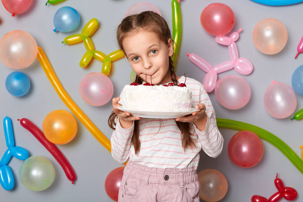 Funny hungry little girl with pigtails standing against gray wall decorated with colorful balloons, holding licking cake at her birthday party. - Zdjęcie, obraz