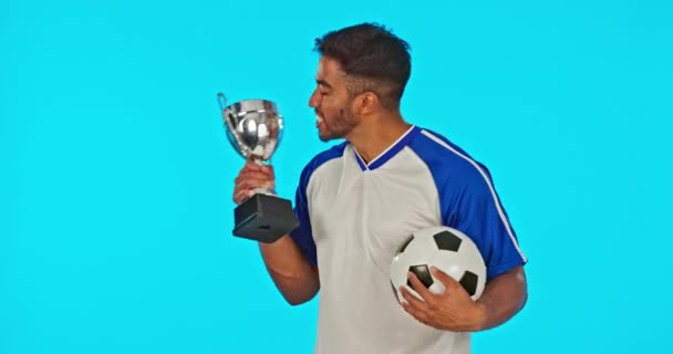 Win, voetbal en gezicht van een man met een trofee geïsoleerd op een blauwe achtergrond in de studio. Gelukkig, succes en portret van een voetballer die een prijs kust voor het winnen van een wedstrijd, prestatie en viering. - Video