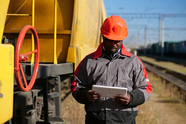 Inspector of wagons at freight train station looks in tablet computer. African american railway man with tablet pc at freight train terminal - Zdjęcie, obraz