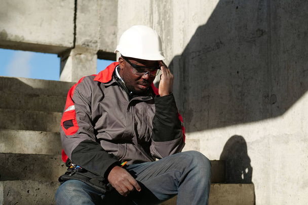 Afrikkalainen amerikkalainen työmies istuu portaissa rakennustyömaalla. Hän ei voi hyvin ja hänellä on päänsärky, hän koskettaa päätään - Valokuva, kuva
