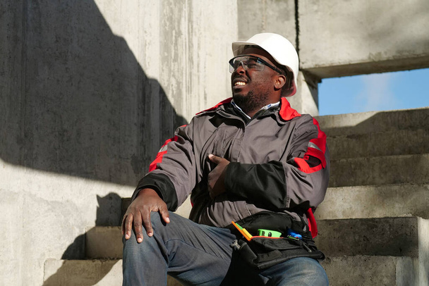 Afrikai amerikai munkás ül a lépcsőn az építkezésen. Nem érzi jól magát, fáj a hasa, megérinti a gyomrát. - Fotó, kép