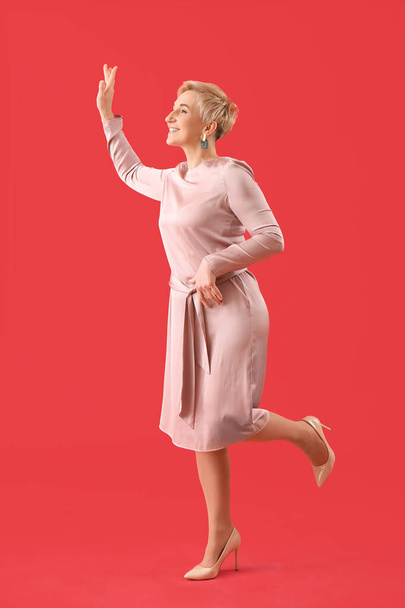 Stylish mature woman waving hand on red background - Фото, изображение