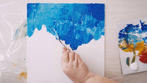 Art studio. Creative process. Abstract artwork. Unrecognizable woman painting with spatula and blue paint on white canvas. - Foto, imagen