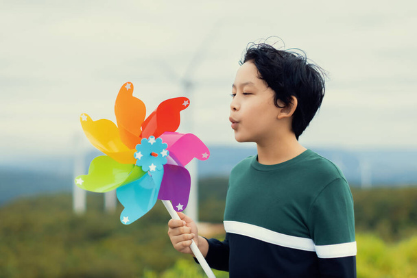 Progressive young asian boy playing with wind pinwheel toy in the wind turbine farm, green field over the hill. Green energy from renewable electric wind generator. Windmill in the countryside concept - Foto, Bild