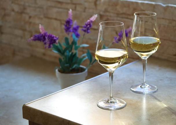 Two glasses of white wine on the kitchen table in front of the window with a purple flower on the back on a sunny day. Transparent glasses of white wine on a table with copy space for text - Фото, зображення
