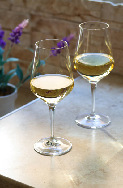 Two glasses of white wine on the kitchen table in front of the window with a purple flower on the back on a sunny day. Close up of a glasses of white wine on a table in natural lights - Photo, Image