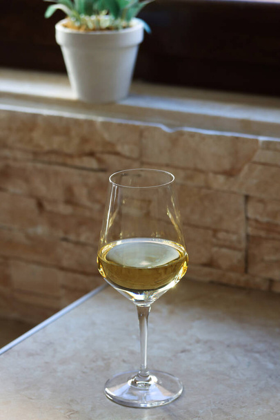 A glass of white wine on the kitchen table in front of the window with a flower on the back on a sunny day. Close up of a glass of white wine on a table with copy space for text - Fotografie, Obrázek