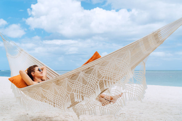 Young woman relaxing in wicker hammock on the sandy beach on Mauritius coast and enjoying wide ocean view waves. Exotic countries vacation and mental health concept image. - Foto, imagen