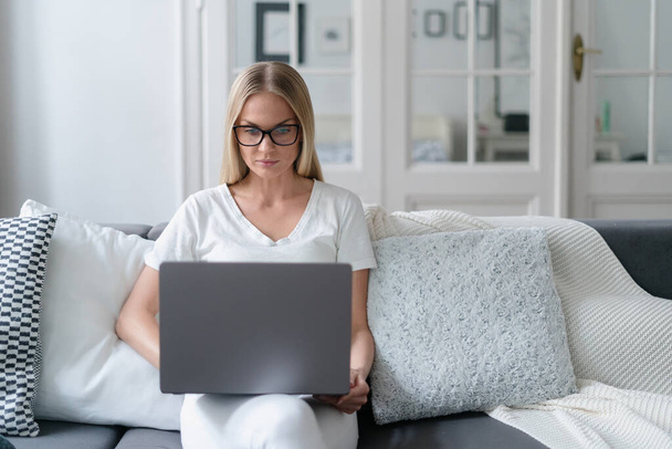 συγκεντρωμένη γυναίκα που εργάζεται από το σπίτι, κάθεται στον καναπέ άνεση, κοιτάζοντας την οθόνη του φορητού υπολογιστή και την αναζήτηση πληροφοριών, μάθηση και online εκπαιδευτικές έννοιες - Φωτογραφία, εικόνα