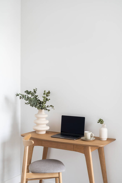 laptop aberto, vaso de cerâmica com ramo de eucalipto e xícara de café em mesa de madeira perto de cadeira confortável em apartamento moderno com parede branca, conceito de escritório em casa - Foto, Imagem