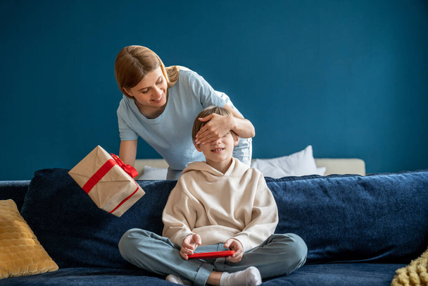 Young happy loving mother closing eyes of child congratulating little boy son with happy birthday at home, smiling mom giving wrapped gift box to kid sitting on sofa with smartphone.  - Foto, imagen