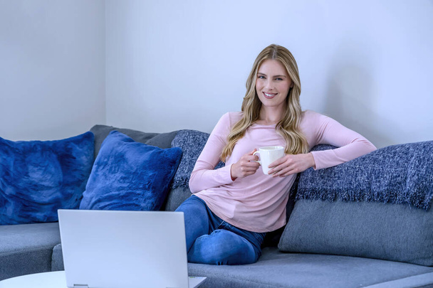 Nette schöne Frau mit blonden Haaren arbeitet am Laptop, während sie sich zu Hause auf das Sofa setzt  - Foto, Bild