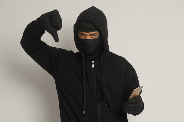 Portré egy titokzatos férfiról, aki fekete kapucnit és maszkot visel, miközben hackeli a mobilját, és egy okostelefont tart a kezében. Izolált kép szürke háttérrel - Fotó, kép