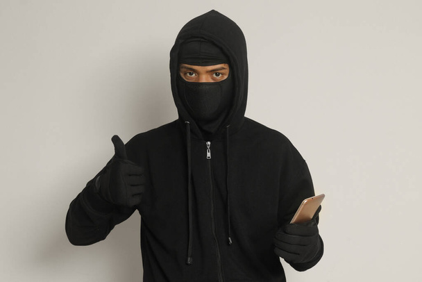 Portrait of mysterious man wearing black hoodie and mask doing hacking activity on mobile phone, hacker holding a smartphone. Isolated image on gray background - Photo, Image