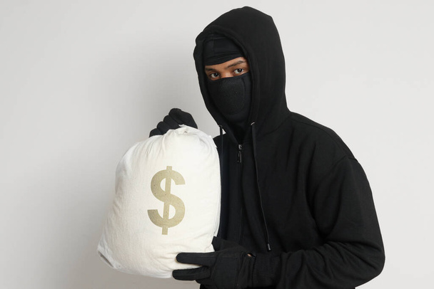 Mysterious robber thief man wearing black hoodie and mask carrying a bag full of money. Isolated image on gray background - Photo, Image