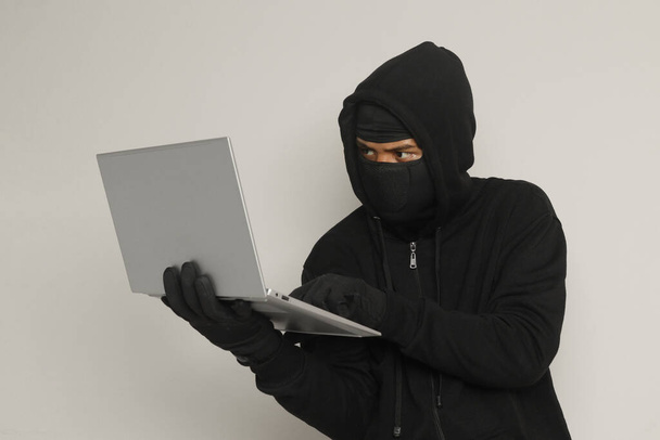 Ritratto di uomo misterioso con cappuccio nero e maschera che fa attività di hacking su laptop, hacker che tiene un personal computer. Concetto di sicurezza informatica. Immagine isolata su sfondo grigio - Foto, immagini