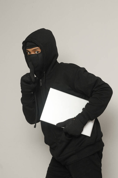 Mysterious robber thief man wearing black hoodie and mask stealing laptop and sneak out. Isolated image on gray background - Photo, Image