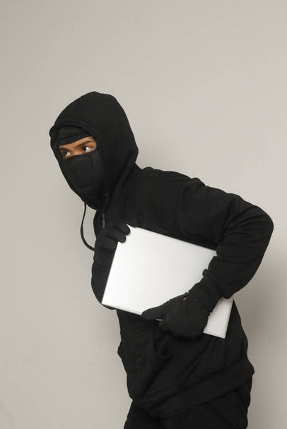 Salaperäinen rosvo varas mies yllään musta huppari ja naamio varastaa kannettava tietokone ja hiipiä ulos. Eristetty kuva harmaalla taustalla - Valokuva, kuva