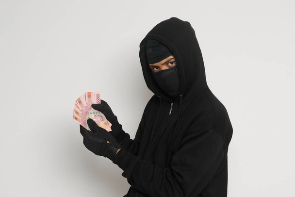 Ritratto di uomo misterioso con cappuccio nero e maschera che ruba centomila rupie con successo alla vittima. Immagine isolata su sfondo grigio - Foto, immagini