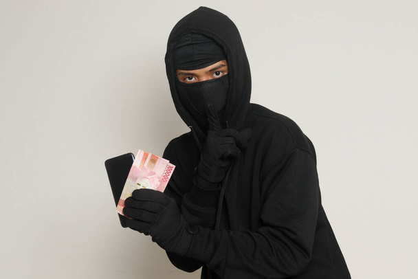 Portrait of mysterious man wearing black hoodie and mask stealing one hundred thousand rupiah successfully from victim. Isolated image on gray background - Photo, Image