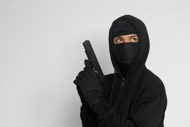 Mysterious man wearing black hoodie and mask holding a pistol, shooting with a gun. Isolated image on gray background - Photo, Image