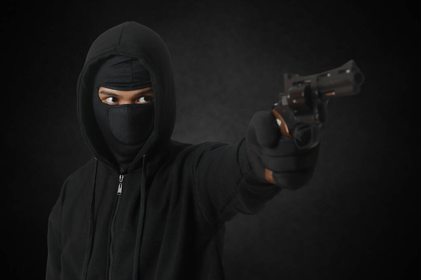 Μυστηριώδης άντρας φοράει μαύρη κουκούλα και μάσκα κρατάει πιστόλι, πυροβολεί με όπλο. Μεμονωμένη εικόνα σε σκοτεινό περιβάλλον - Φωτογραφία, εικόνα