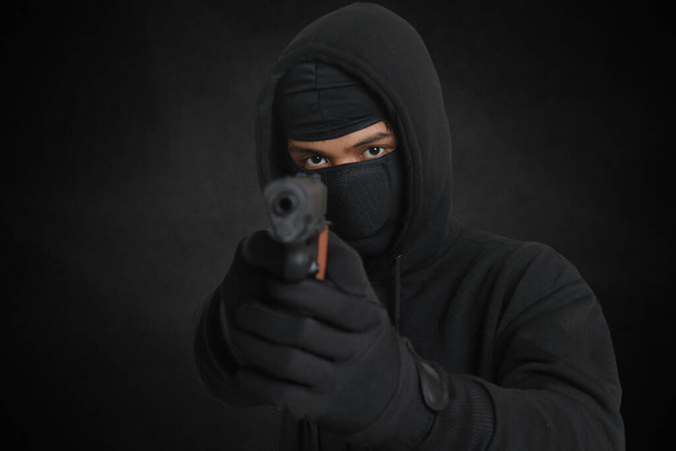Hombre misterioso con capucha negra y máscara sosteniendo una pistola, disparando con un arma. Imagen aislada sobre fondo ambiente oscuro - Foto, Imagen